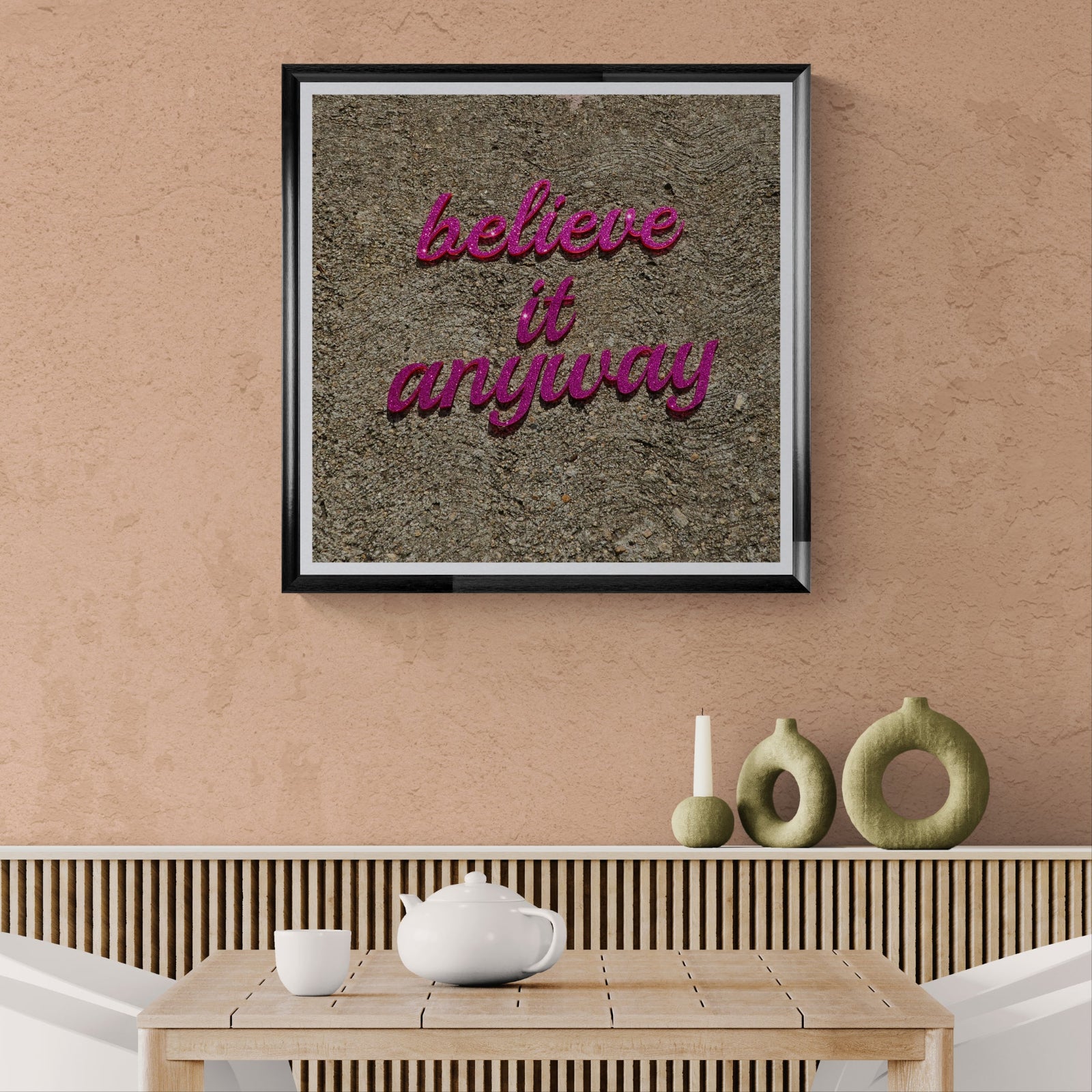 Believe It Anyway magenta typography art print displayed as 30 × 30 wall art in a black frame above a natural wood dining table on a warm peach wall.