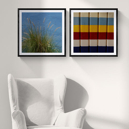 Two 20x20 matte art prints featuring ornamental grass against a blue wall and colorful geometric painted stripes, displayed in black frames above a white armchair.