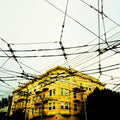 Electric Veins film photograph of a yellow apartment building beneath a dense network of overhead transit wires, creating a graphic urban pattern against a pale sky.