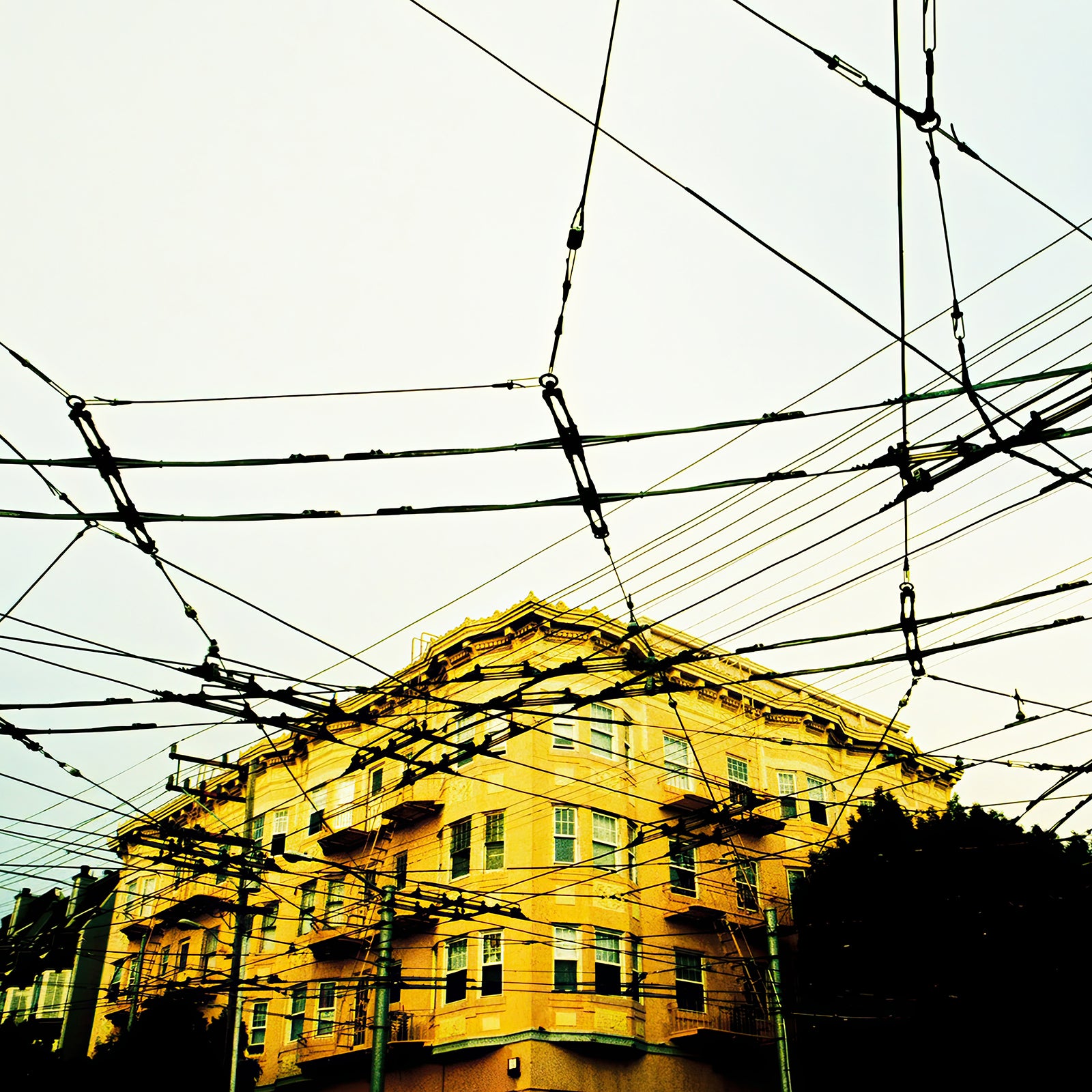 Electric Veins film photograph of a yellow apartment building beneath a dense network of overhead transit wires, creating a graphic urban pattern against a pale sky.