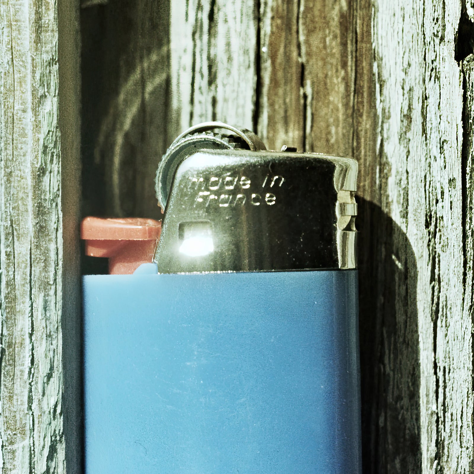 French Spark film photograph of a blue disposable lighter tucked between weathered wooden boards, with “Made in France” visible on the metal top.