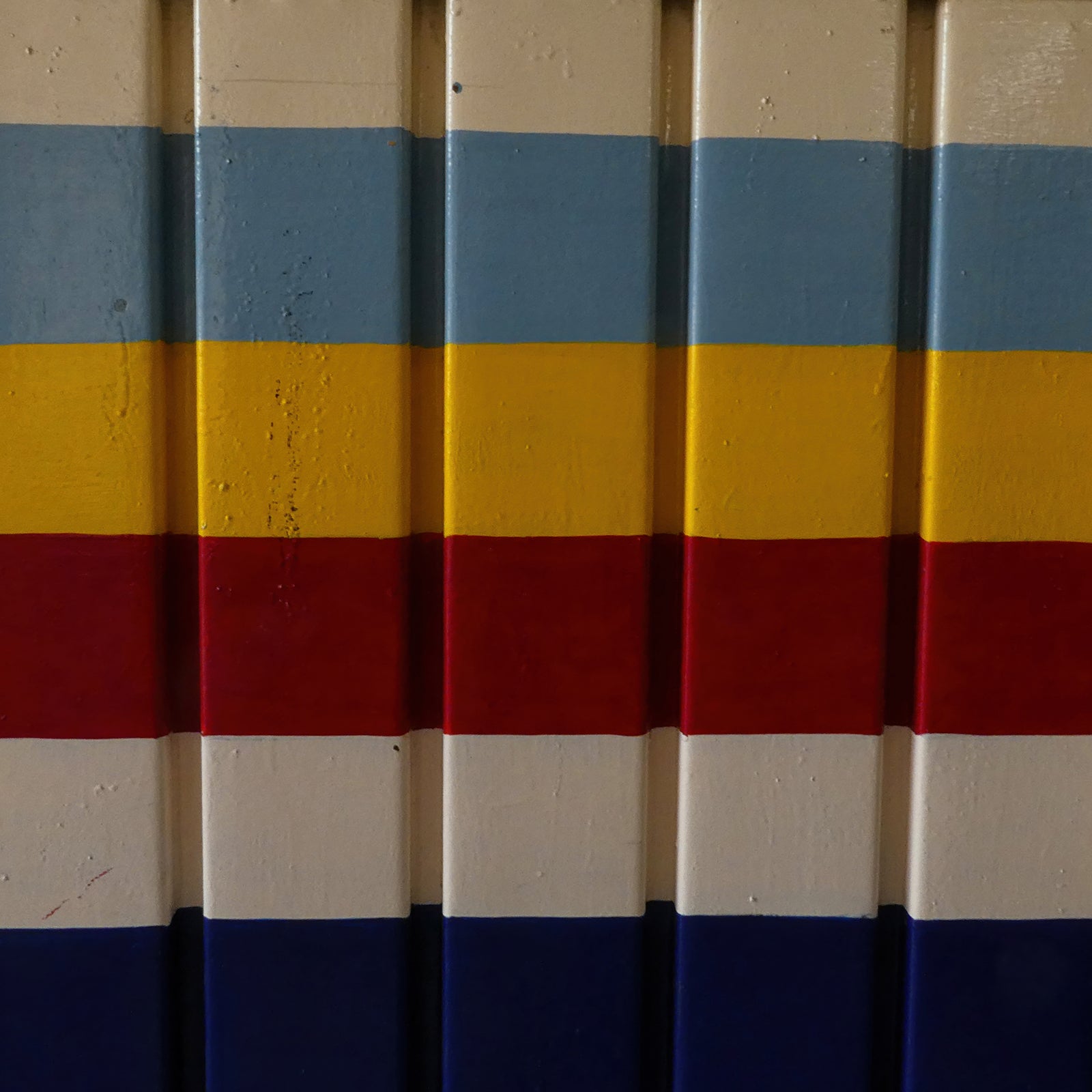 Colorful horizontal bands of blue, yellow, red and cream painted across a vertically corrugated architectural surface.